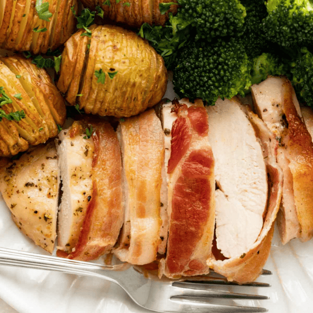 baked chicken wrapped in bacon served with potatoes and broccoli on a white plate with a fork
