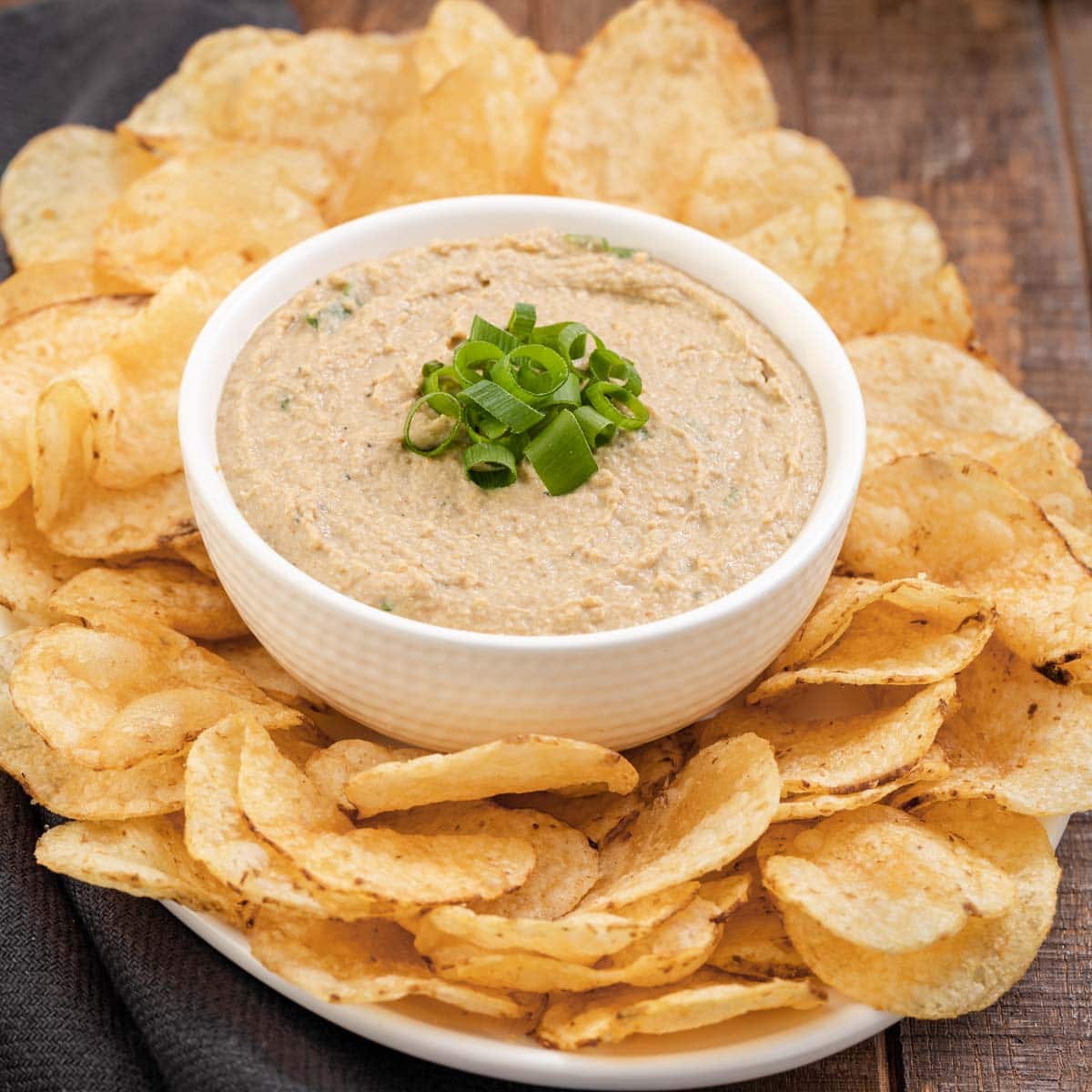 Clam Dip finished in bowl with green onions on top and bowl surrounded by potato chips on platter