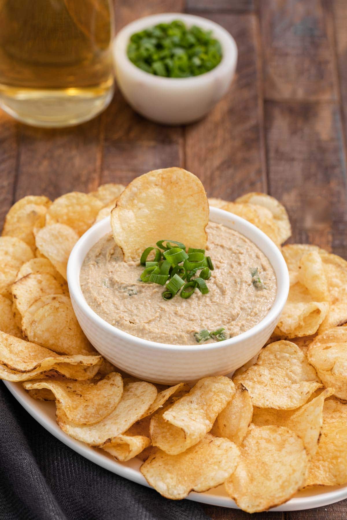 Clam Dip finished in bowl with green onions and chip on top and bowl surrounded by potato chips on platter