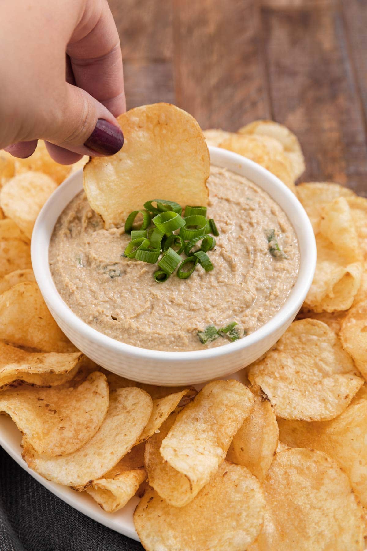 Clam Dip finished in bowl with green onions on top and bowl surrounded by potato chips on platter, dipping chip into dip