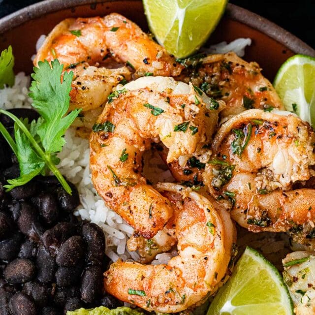 Shrimp finished in a bowl with lime wedges, black beans, rice, and avocado close up