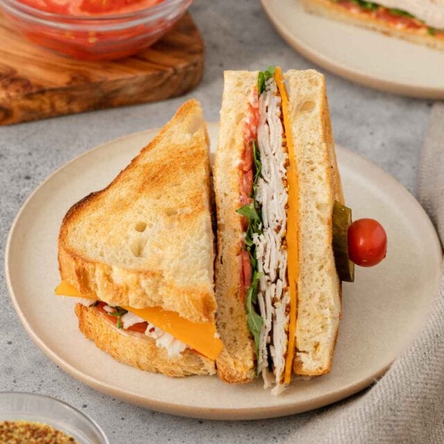 Turkey Sandwich halves on plate. One half laying flavor, other half propped up on flat half. Propped half held together with small tomato and pickle slice on toothpick. 1x1