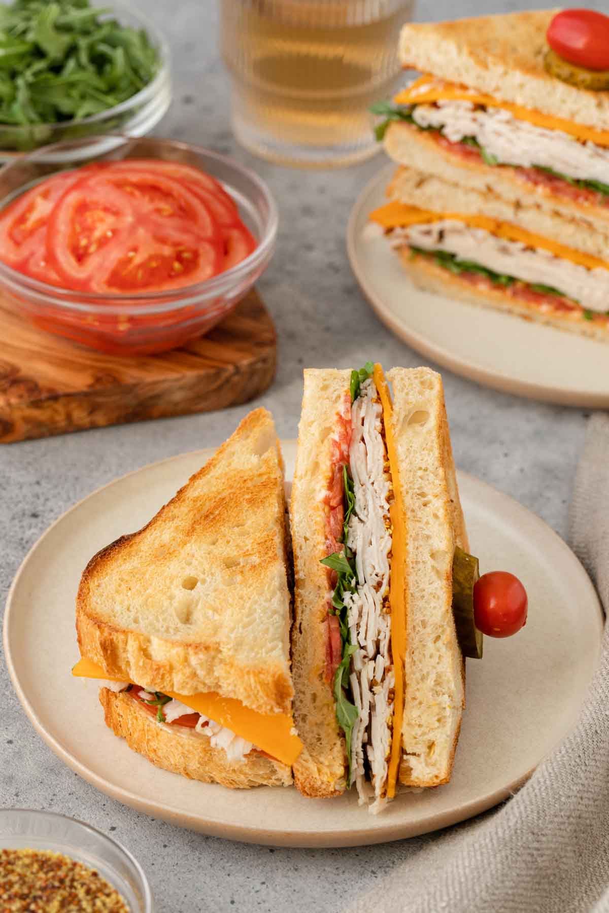 Turkey Sandwich halves on plate. One half laying flavor, other half propped up on flat half. More halves stacked on plate and bowl of tomatoes in background.