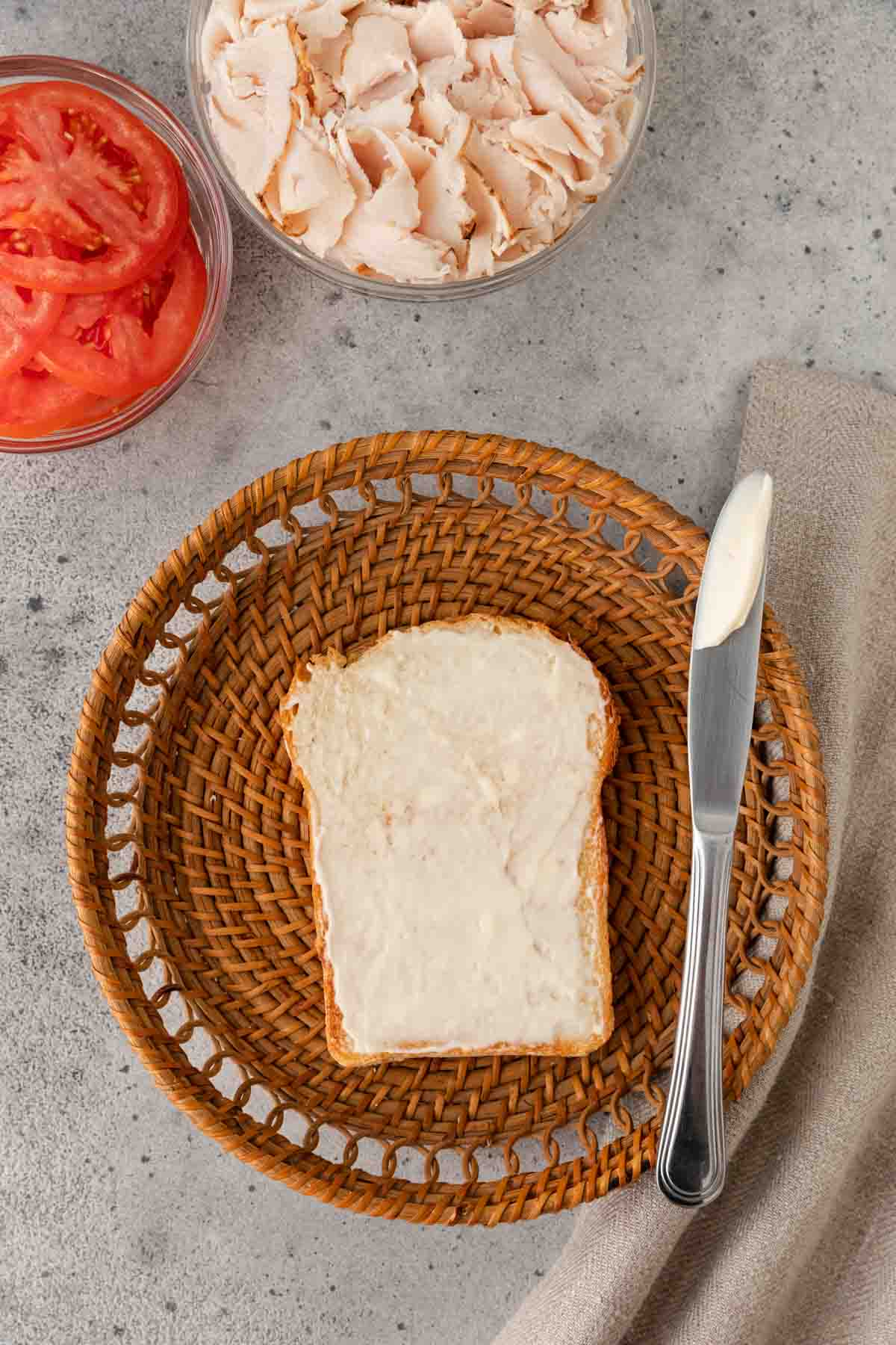 Turkey Sandwich slice of bread on plate with mayo speed on top with knife resting on plate. Turkey and tomatoes in separate prep bowls at top of frame.