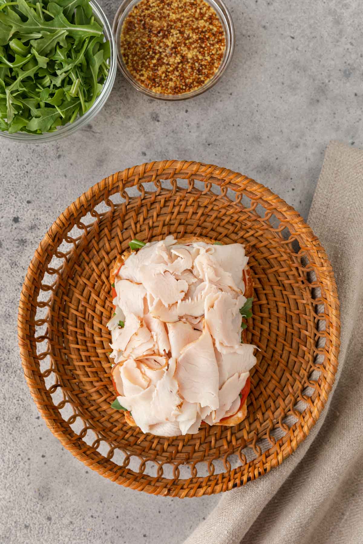 Turkey Sandwich slice of bread topped turkey on brown plate. Arugula and whole grain mustard in prep bowls in top left of frame