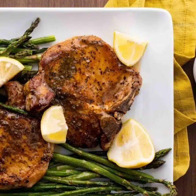 Pork Chops on a square plate with asparagus and lemon wedge garnishes