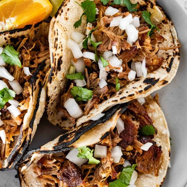 Instant Pot Pork Carnitas plated in charred flour tortillas with onions and cilantro on top, garnished with lime and orange wedges