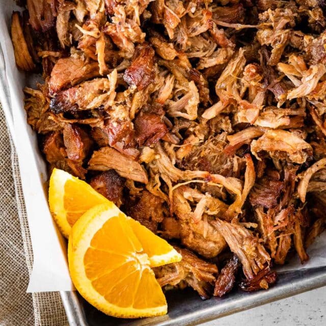 Instant Pot Pork Carnitas shredded meat on baking sheet after roasting. garnished with orange slices, 1x1