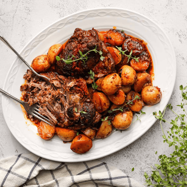 Lamb Served On White Platter With Carrots And Potatoes With Serving Utensils