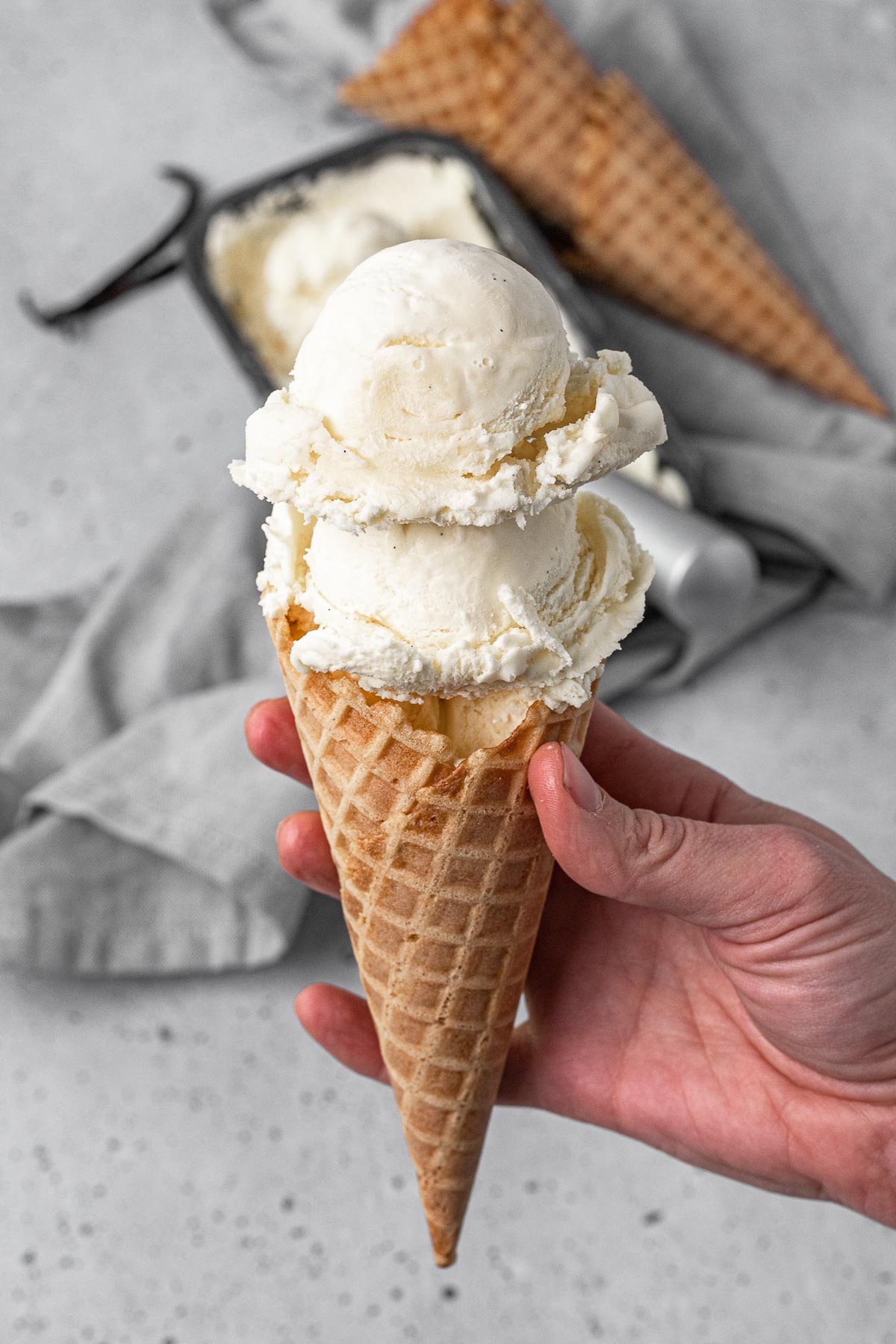 French Vanilla Ice Cream hand holding waffle cone with two scoops of ice cream and ice cream cones in background