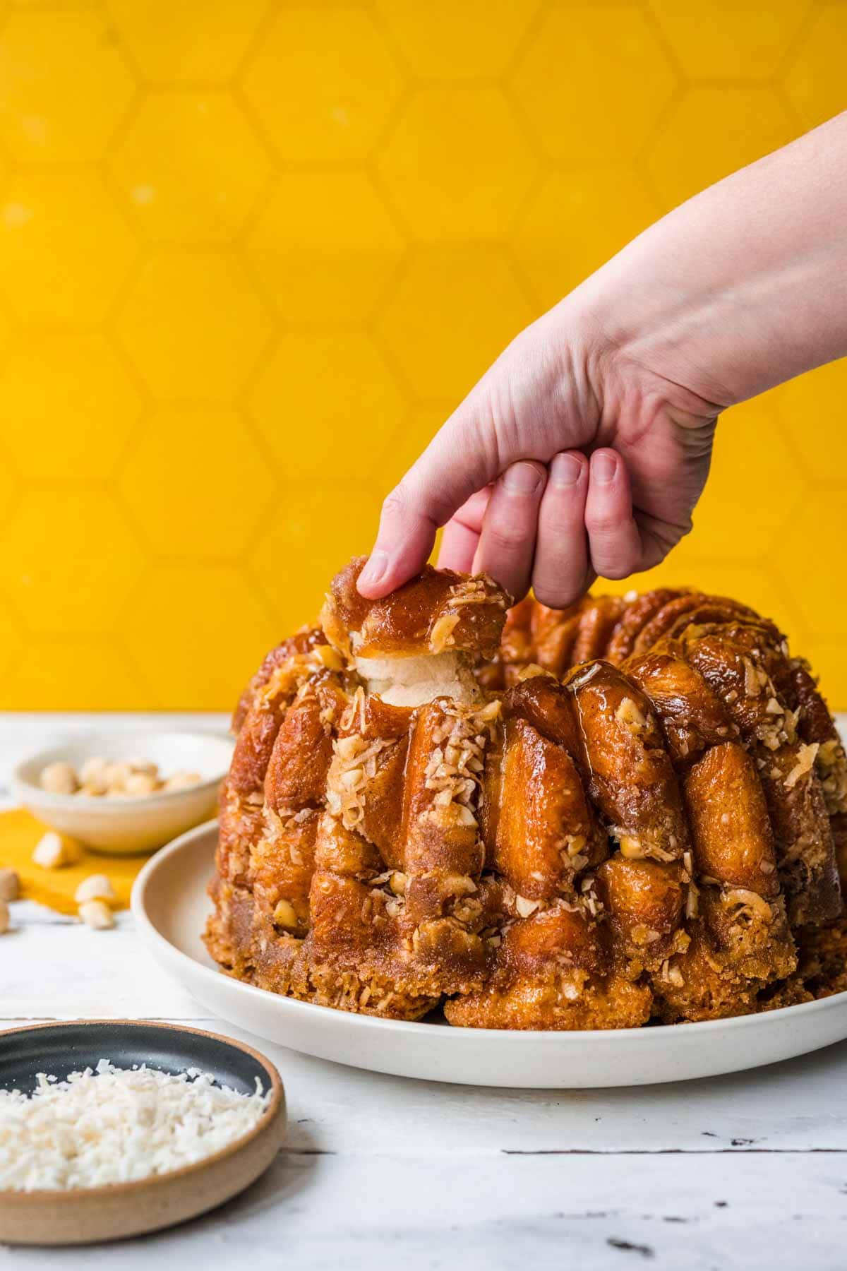 Hawaiian Monkey Bread finished on plate with coconut shreds and macadamia nuts in small bowls next to plate, hand pulling piece off top