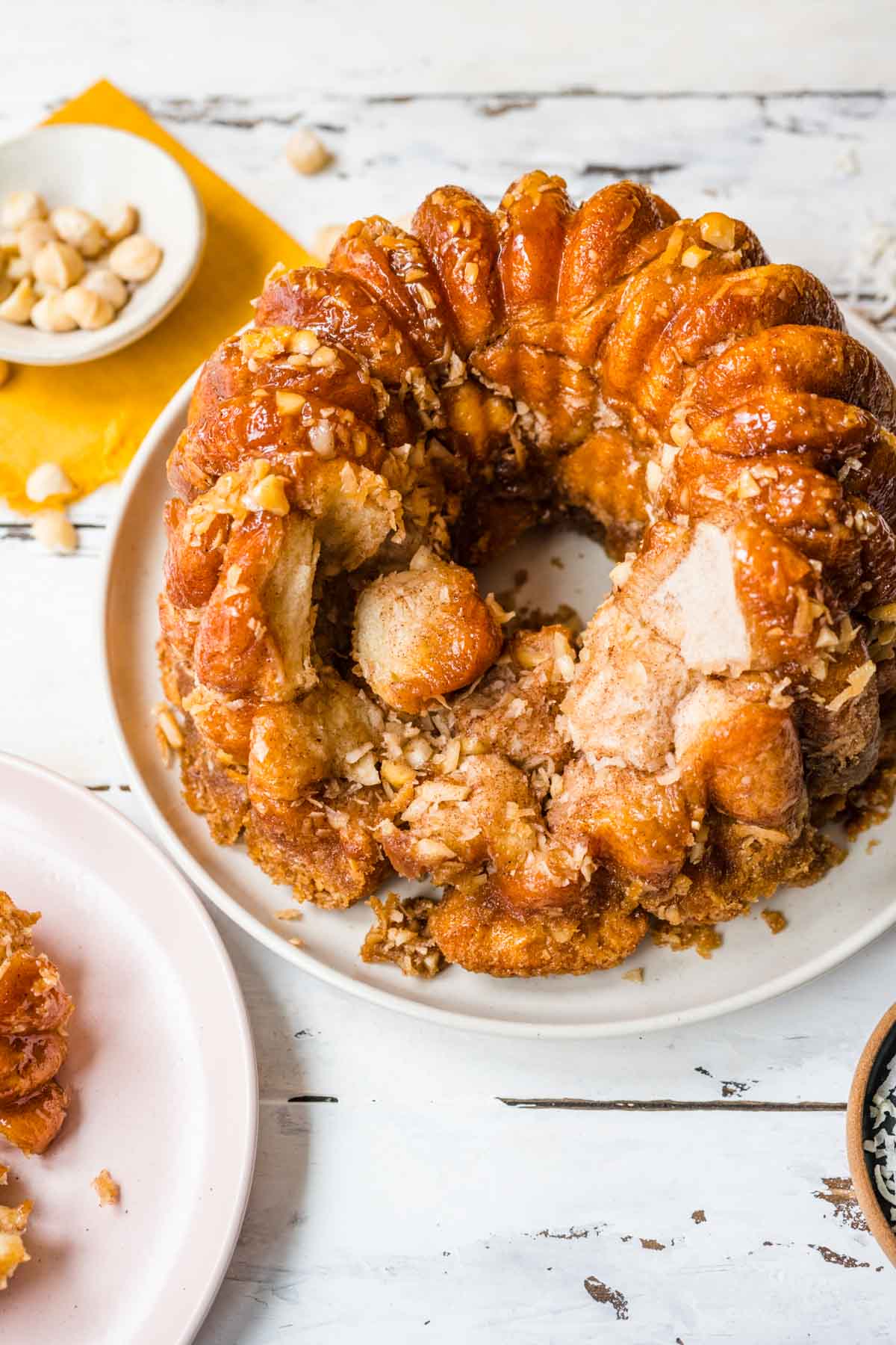 Hawaiian Monkey Bread finished on plate with pieces missing and coconut shreds and macadamia nuts in small bowls next to plate, top down view