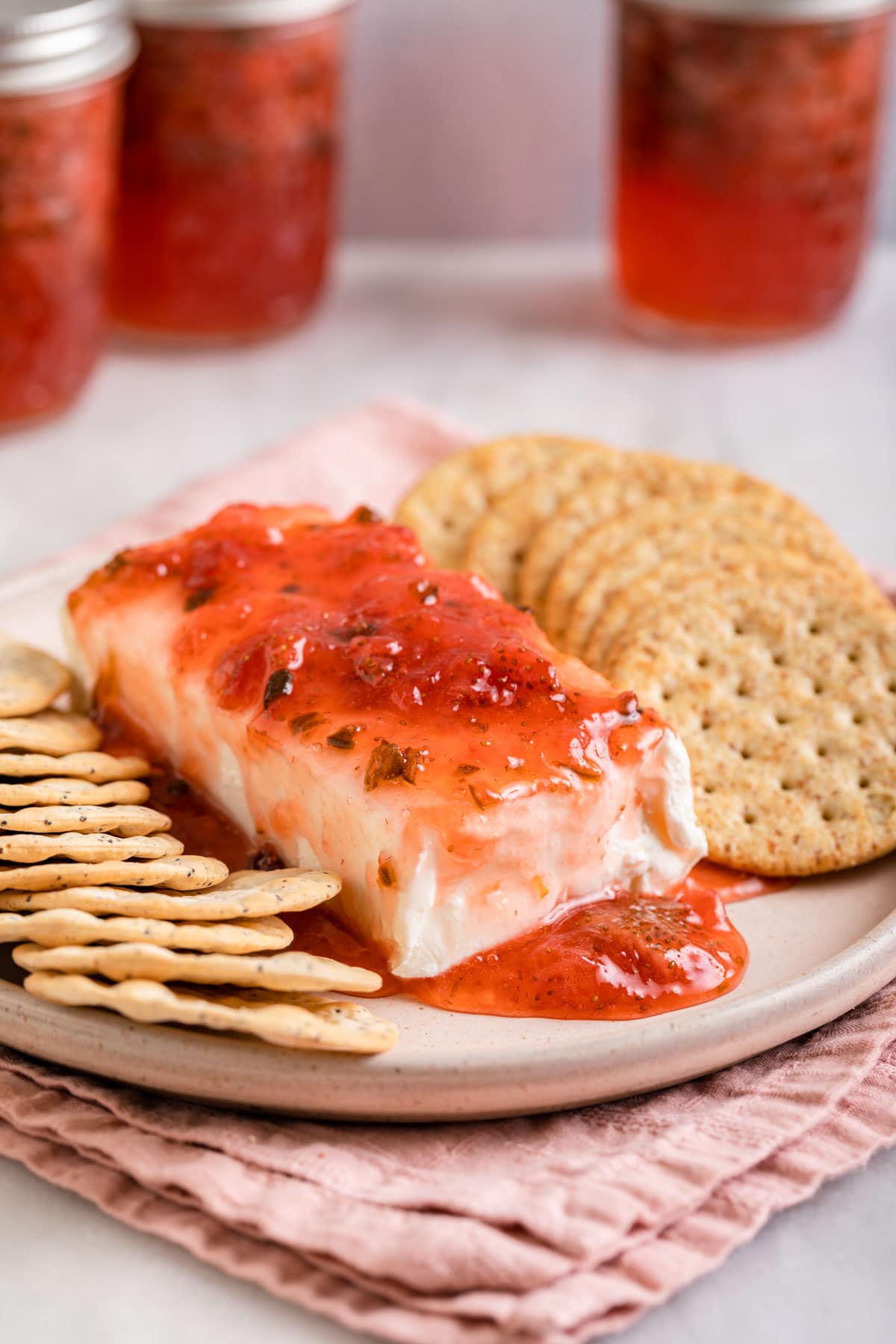 Strawberry Jalapeno Jam poured on block of cream cheese with round crackers on plate with jam jars in background