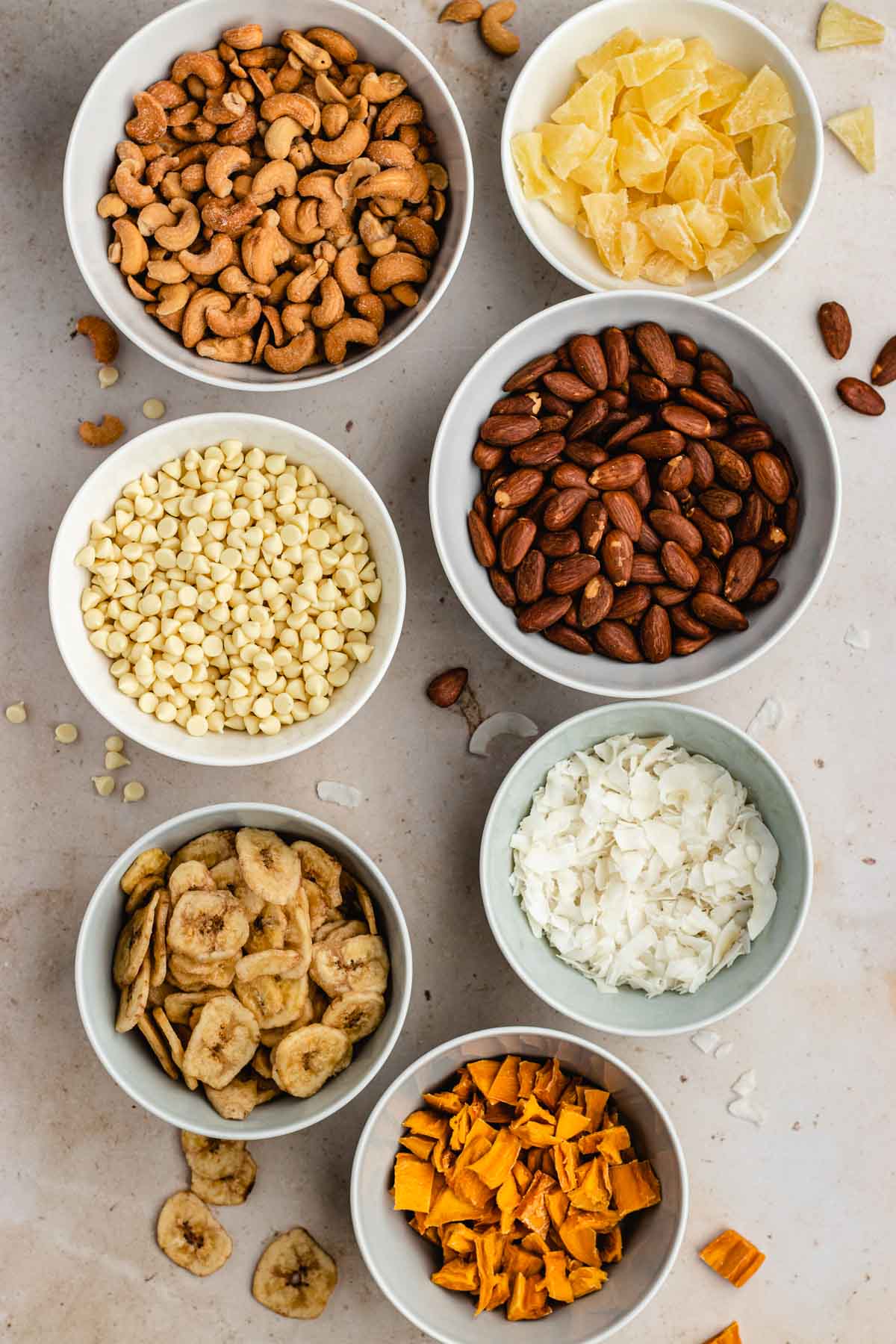 Tropical Trail Mix ingredients in separate small bowls. Two bowls per row going left to right it' is cashews, pineapple, white chocolate, almonds, banana chips, coconut, and mango on its own row.