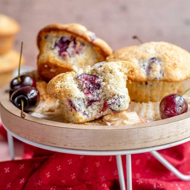 Cherry Muffins piled on cake stand with front muffin having bite taken out, 1x1