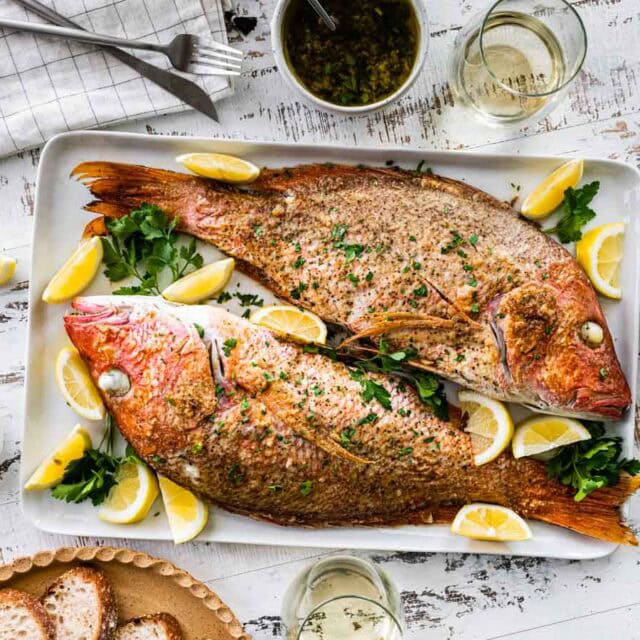 Two whole baked red snappers on serving dish with lemon wedges.
