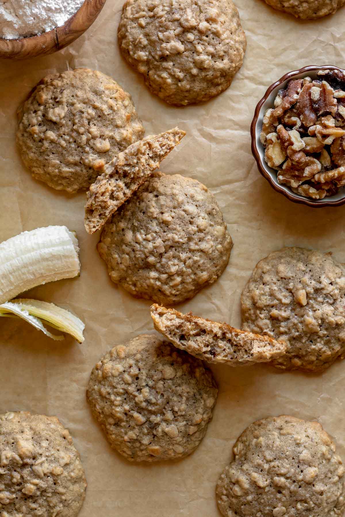 Banana Bread Cookies some cookies on brown parchment paper with half peeled banana off to side and one cookie broken in half, plus small bowl of walnuts on right