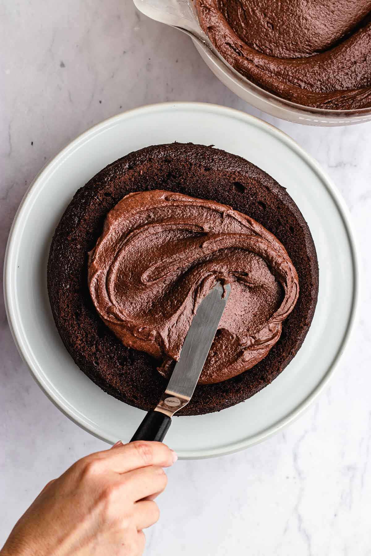 Dark Chocolate Frosting spreading on top of chocolate cake on white cake stand