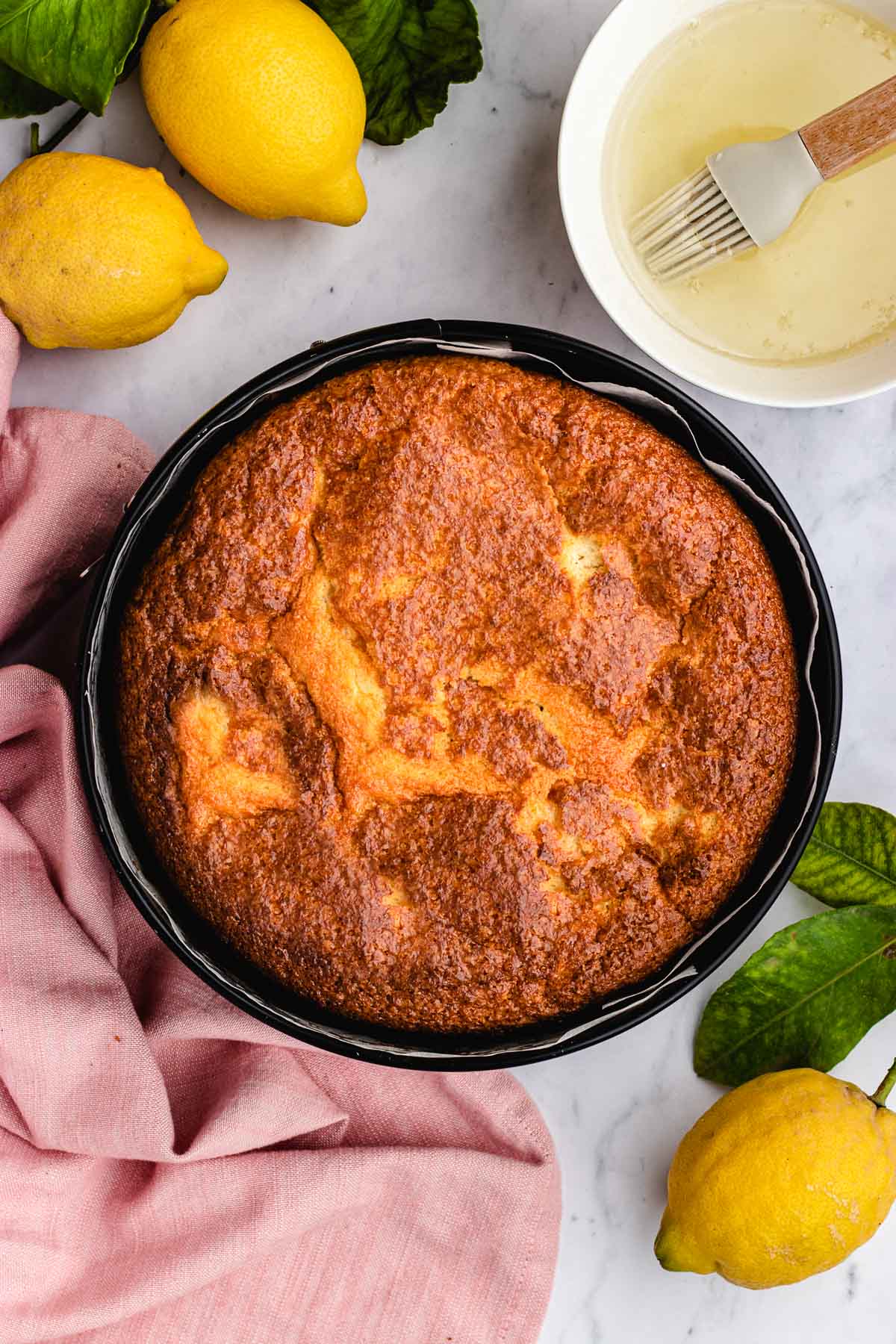 Lemon Olive Oil Cake baked in cake pan. Simple syrup with pastry brush in small bowl in top right corner. Whole lemons around cake pan.