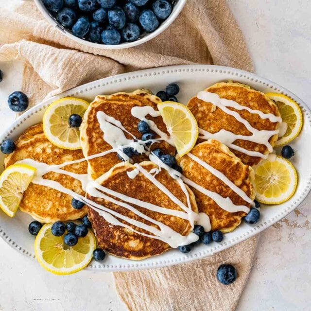 Lemon Ricotta Pancakes cooked pancakes spread and piles on plate with blueberries and lemon wheels, with glaze drizzle, 1x1