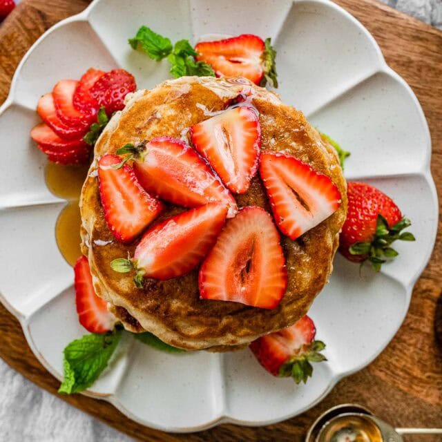 Stack of pancakes on a plate with maple syrup and halved berries.