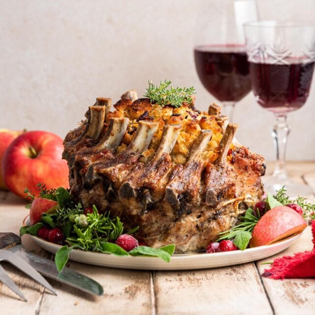 Crown Pork Roast whole roast finished on plate with garnishes around bottom and glasses of red wine in background, 1x1