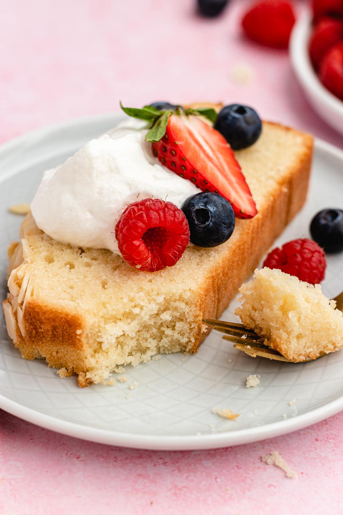 Almond Pound Cake slice on plate with whipped cream and berries on top and fork with bite.