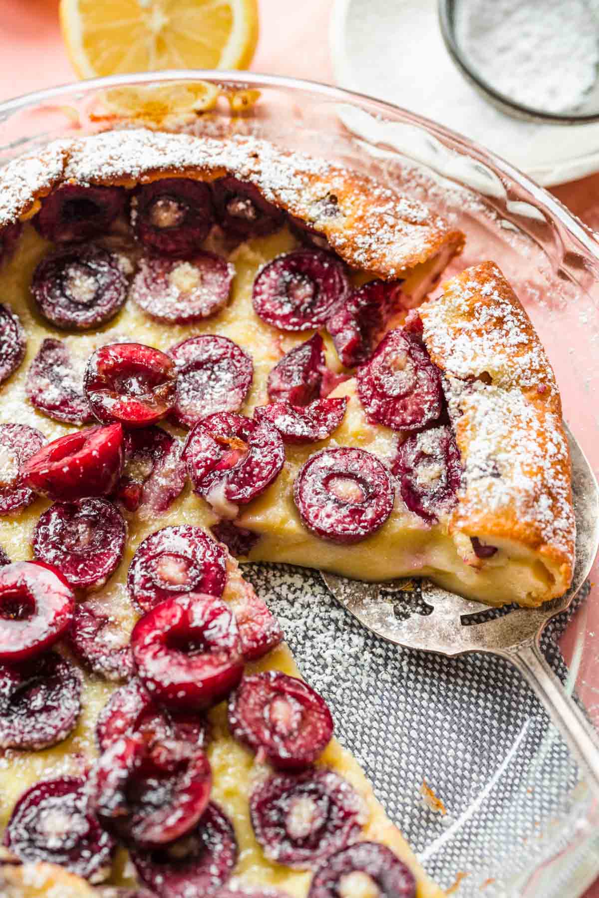 Cherry Clafoutis baked in dish with slice removed and serving spatula under another slice, with pink background and ingredient decorations