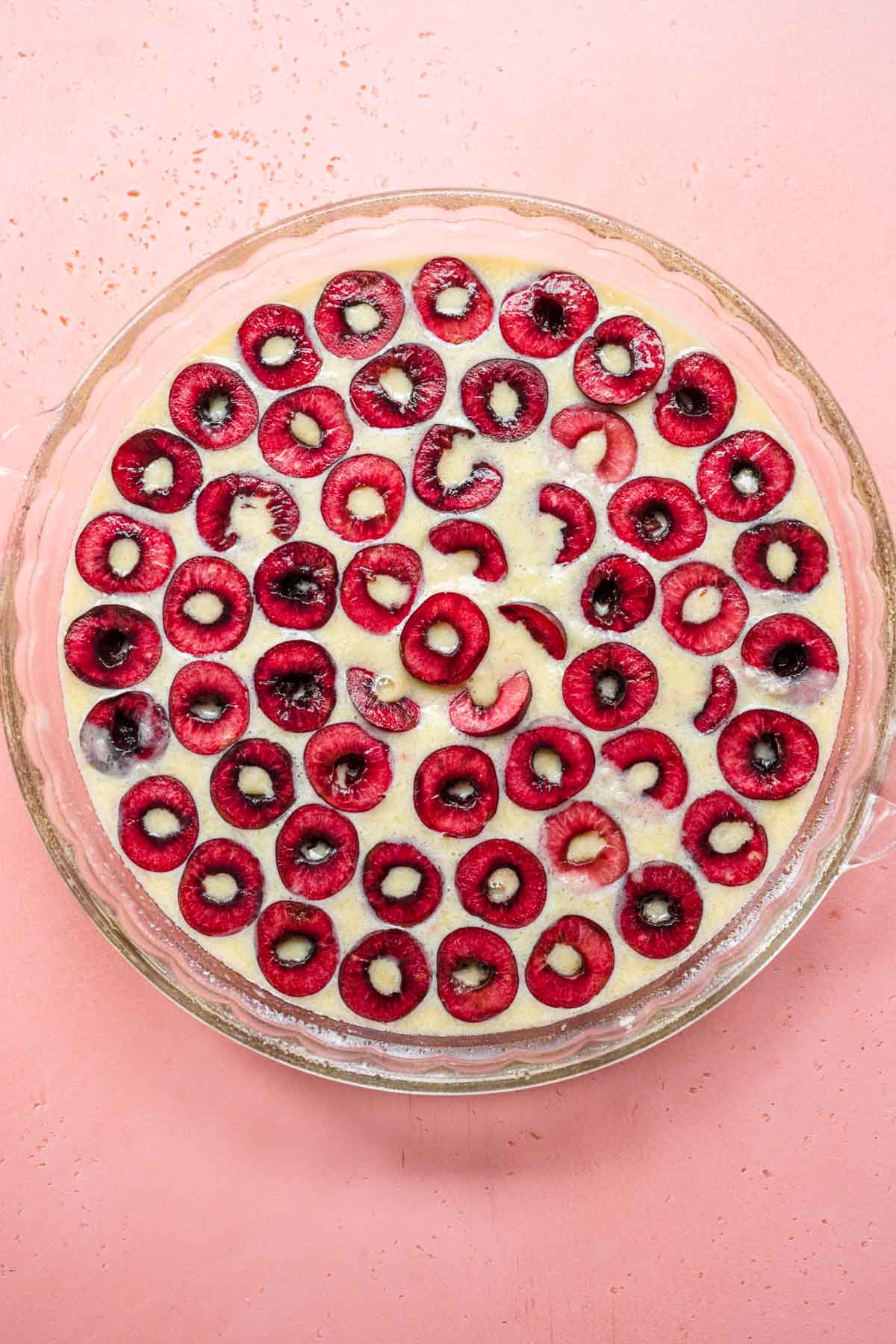 Cherry Clafoutis batter and cherries in glass pie dish before baking, pink background
