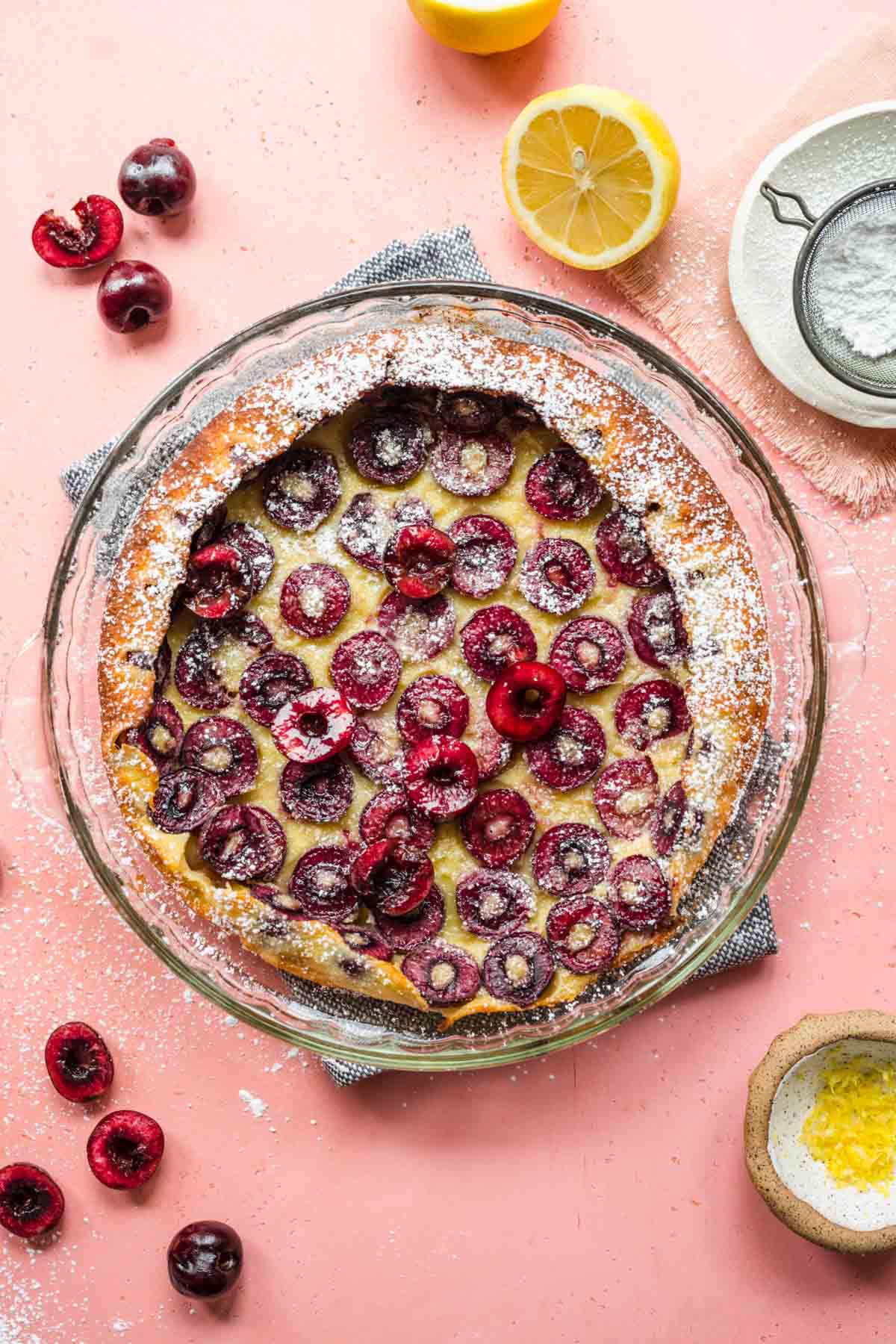 Cherry Clafoutis baked in dish with lemons and cherries surrounding the dish, pink background with ingredient decorations