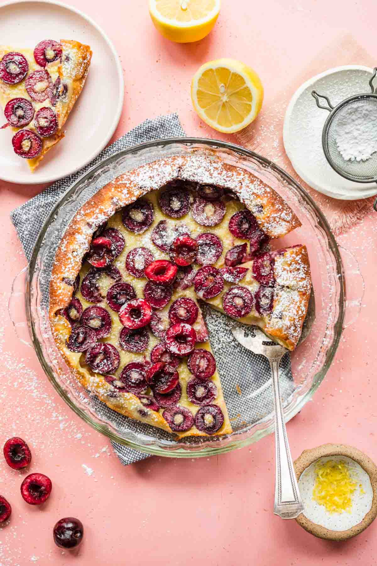 Cherry Clafoutis baked in dish with slice removed and serving spatula under another slice, with pink background and ingredient decorations