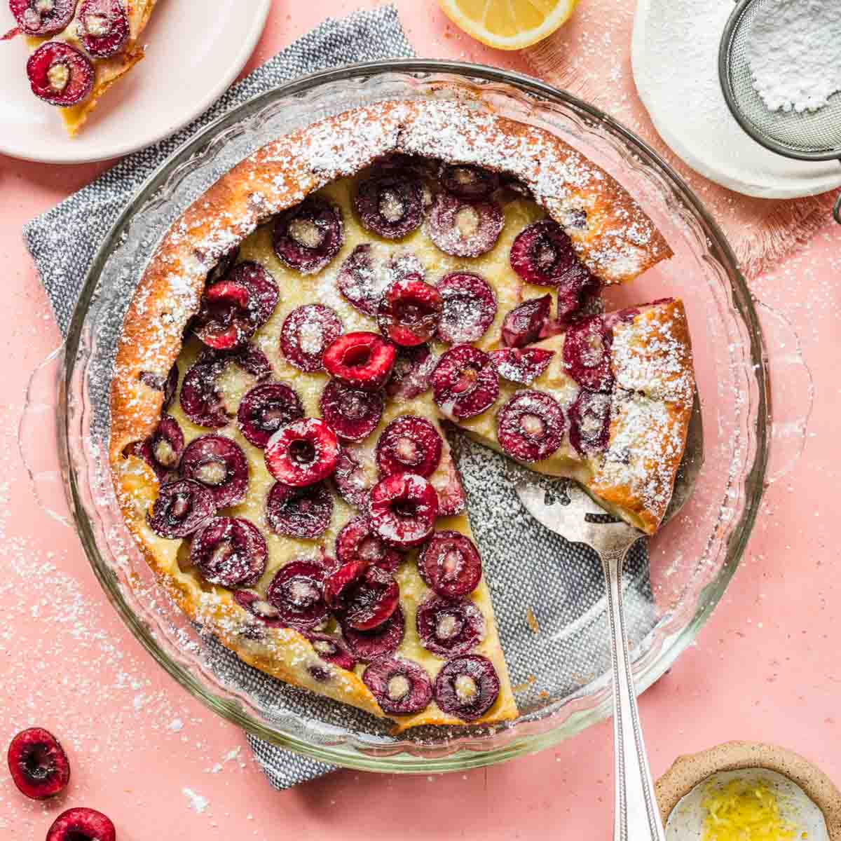 Cherry Clafoutis baked in dish with slice removed and serving spatula under another slice, with pink background and ingredient decorations