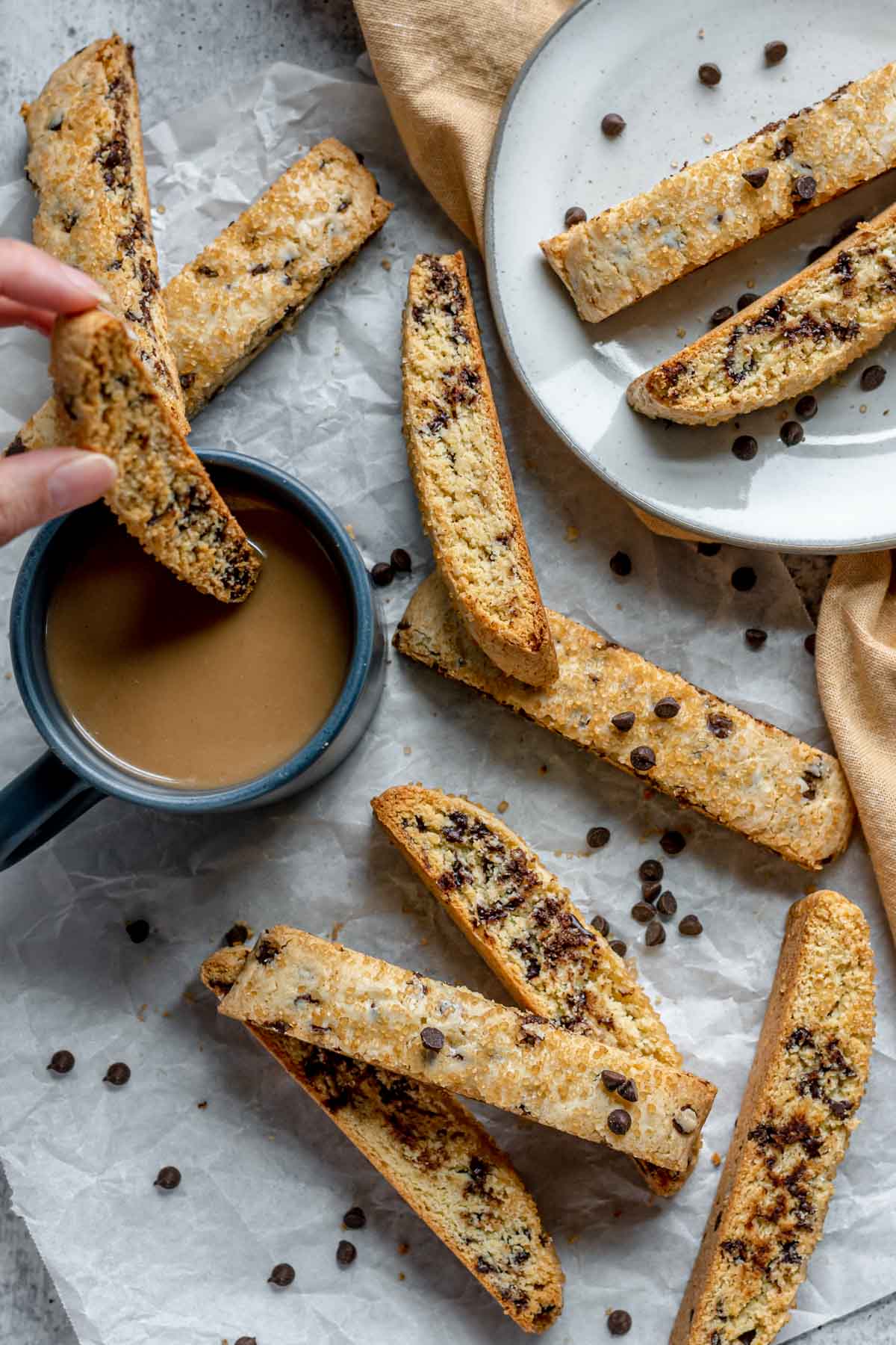 Chocolate Chip Biscotti baked cookie being dunked into cup of coffee with other cookies scattered on counter and plate