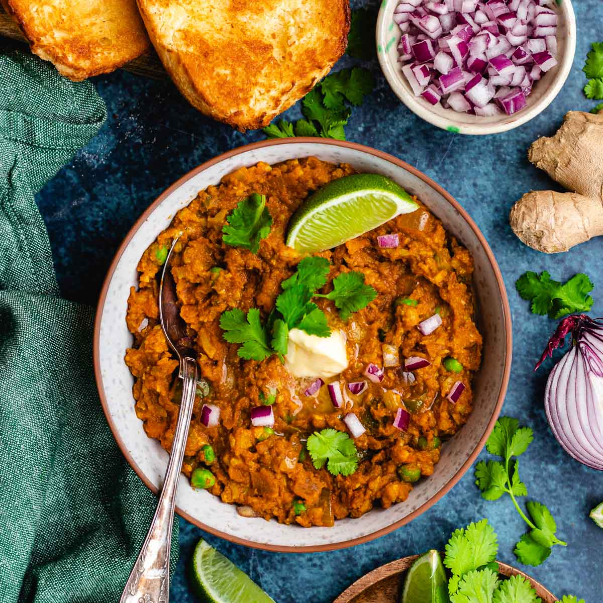 Pav Bhaji cooked and garnished in bowl with spoon with bread and garnishes off to side