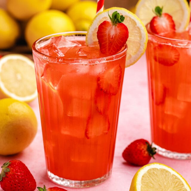 Strawberry Lemonade in glass on pink surface with halved berry and lemon wheel on rim of glass. lemons and berries in background. 1x1
