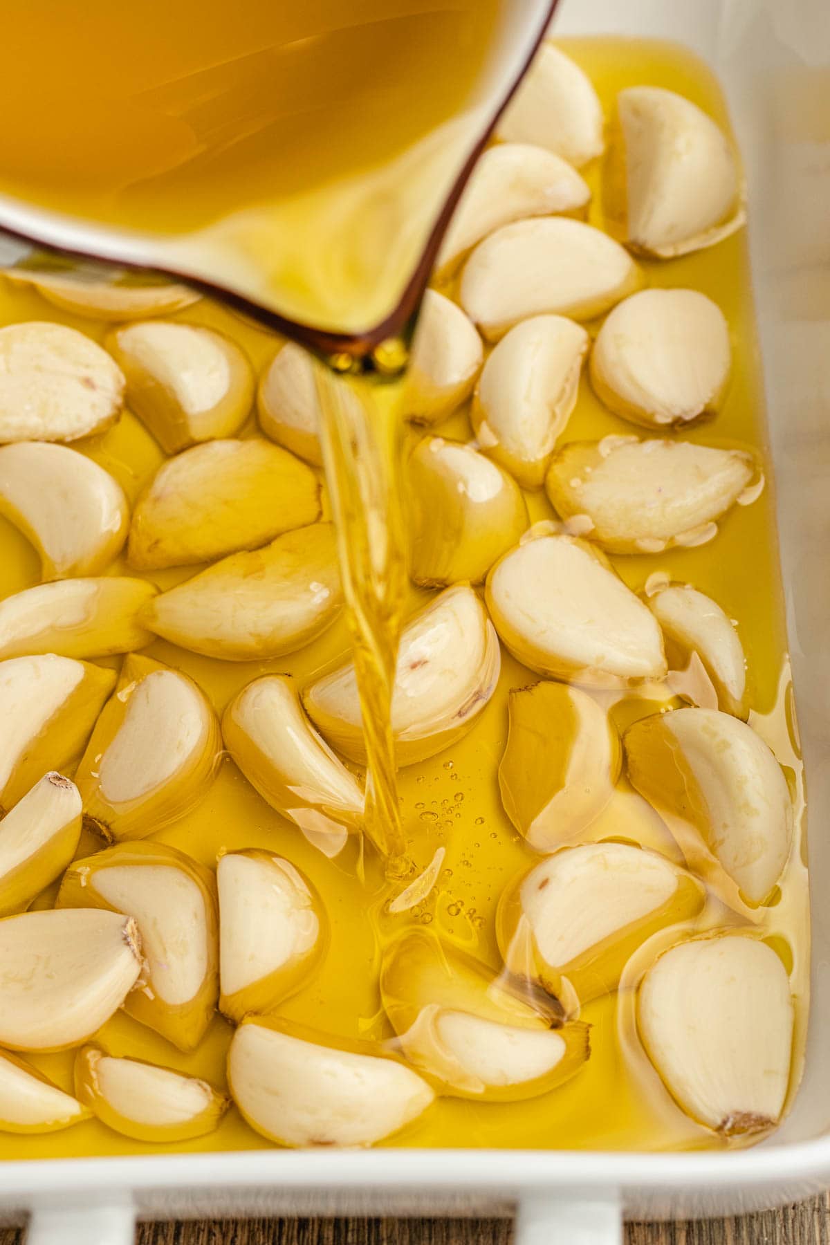 Garlic Confit pouring oil over raw cloves in baking dish