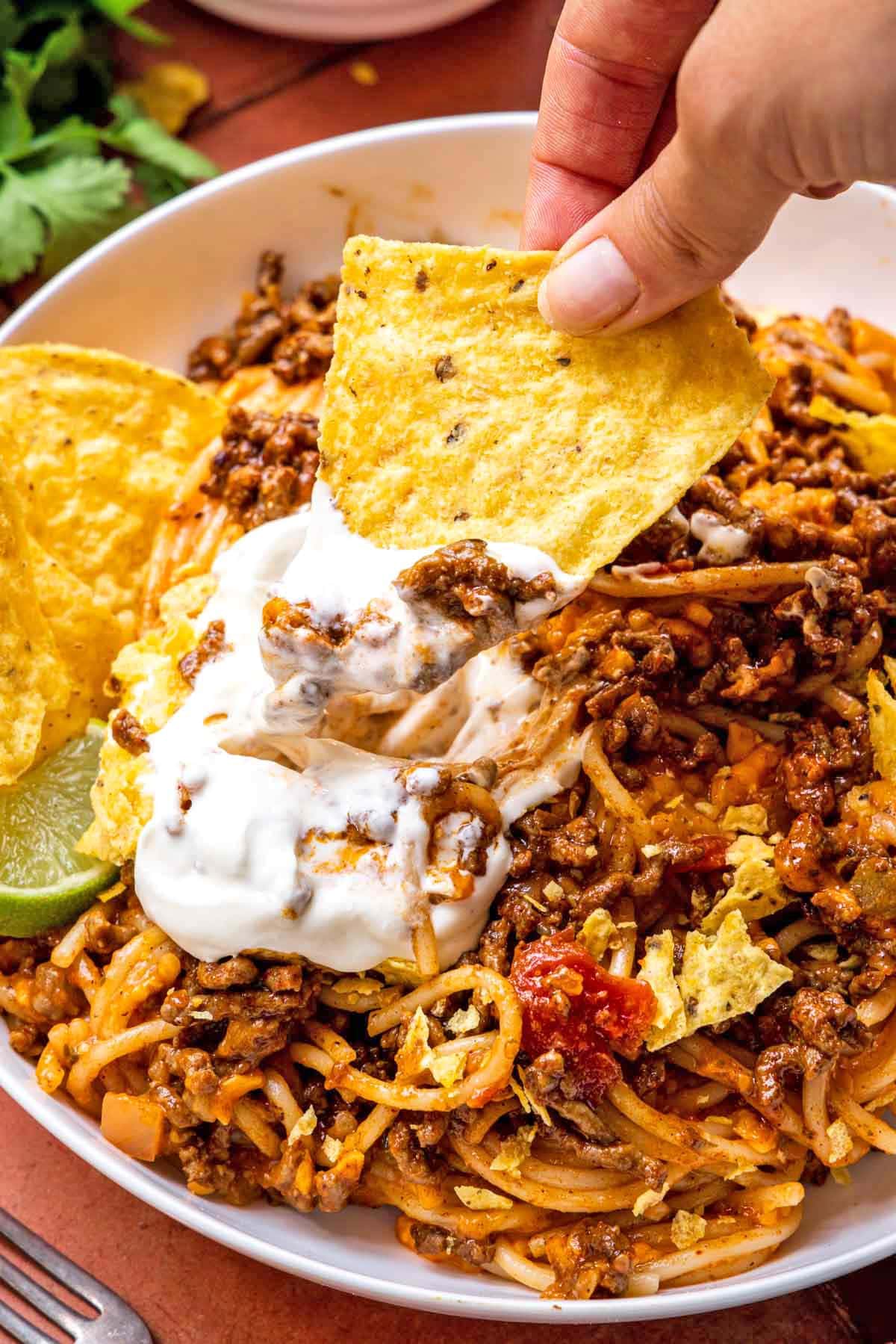 Taco Spaghetti finished pasta dish in bowl with chips, lime, and sour cream garnish, chip scooping some sour cream and sauce