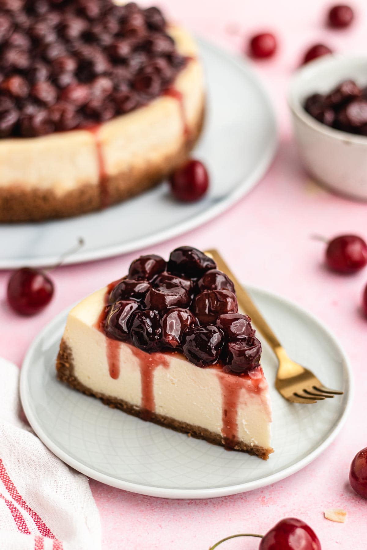 Cherry Cheesecake slice on plate with fork and cheesecake in background
