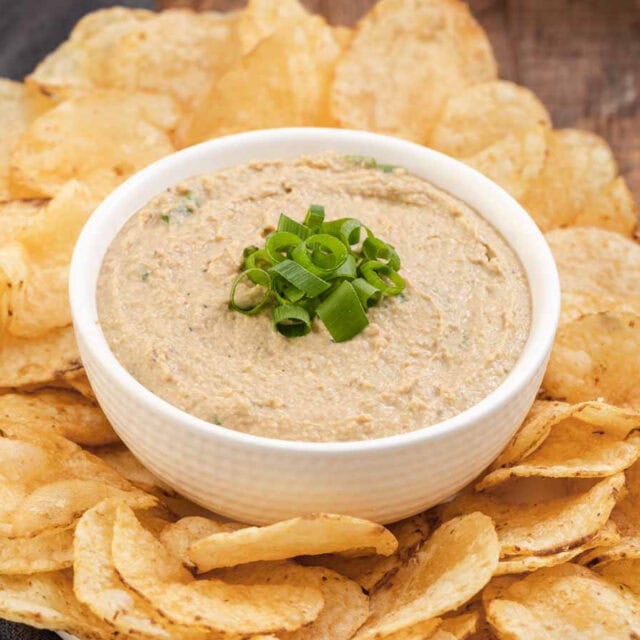 Dip finished in a bowl with green onions on top and bowl surrounded by potato chips on platter, 4x3