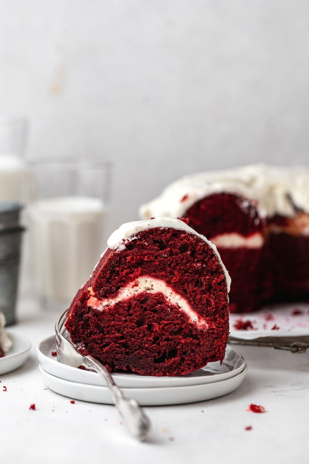 Red Velvet Cheesecake Bundt Cake frosted cake slice on plate with cake in background