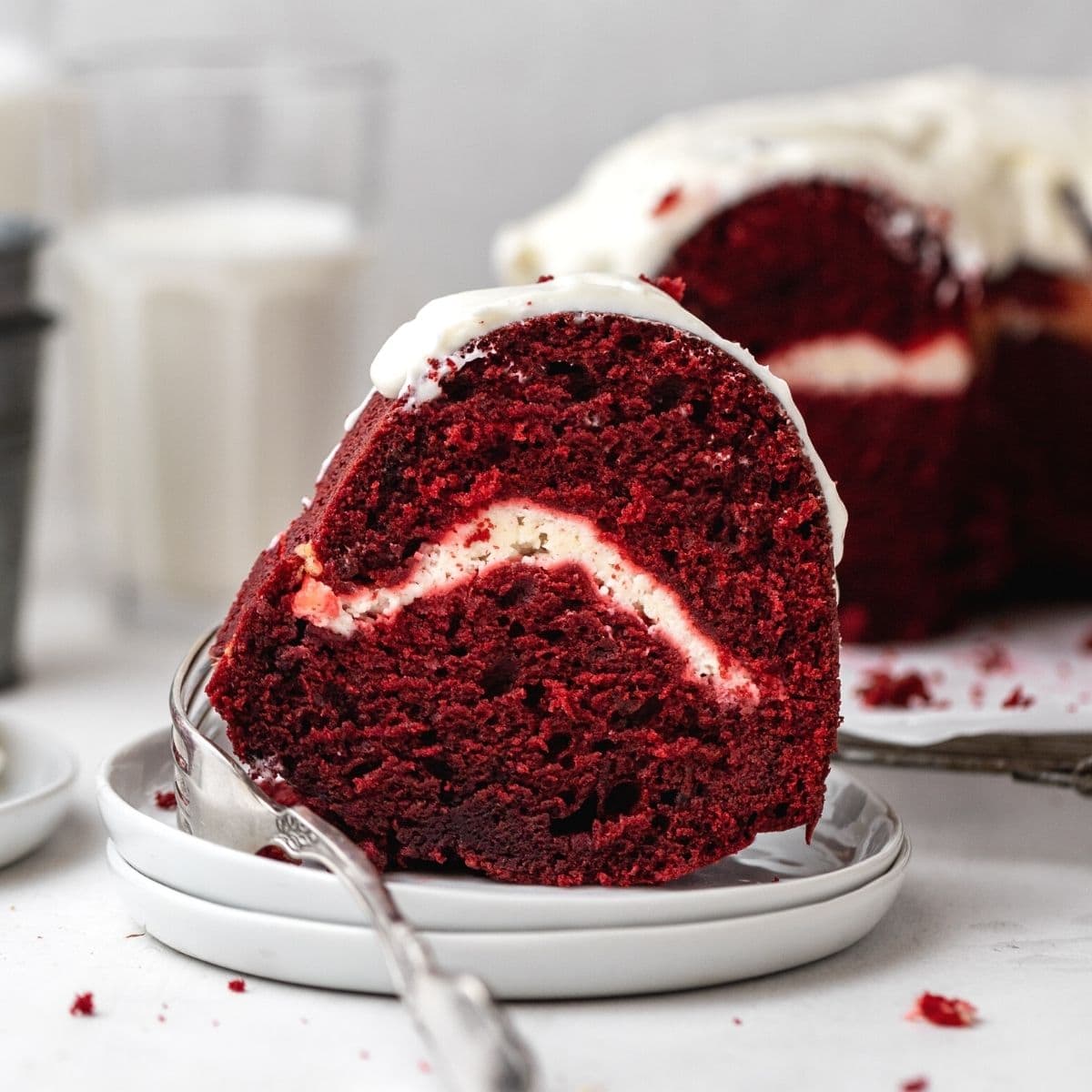 Red Velvet Cheesecake Bundt Cake frosted cake slice on plate with cake in background