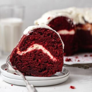 Red Velvet Cheesecake Bundt Cake frosted cake slice on plate with cake in background