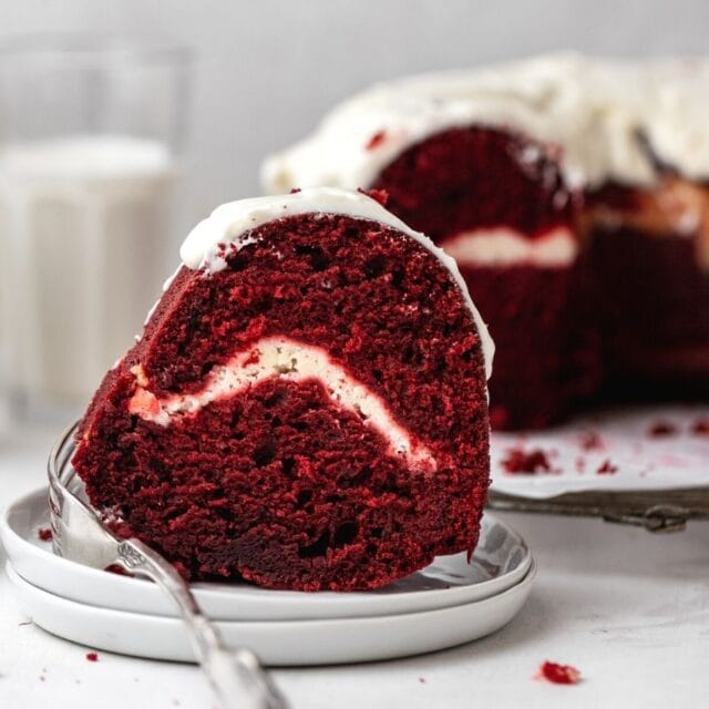 Red Velvet Cheesecake Bundt Cake frosted cake slice on plate with cake in background
