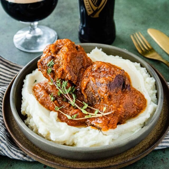 Guinness Braised Short Ribs close up of ribs plated on mashed potatoes, glass of beer and beer bottle slightly in background, 1x1 cropped image
