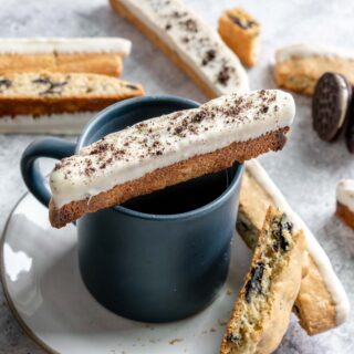 Oreo Dipped Biscotti one biscotti dipped side up resting on coffee cup on plate with more biscotti arranged on and around plate. 1x1