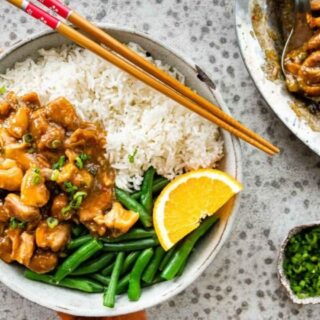 Chicken on a white plate with green beans, rice, an orange slice and chopsticks.