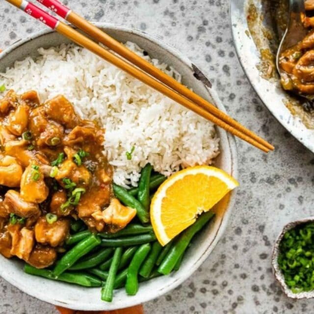 Chicken on a white plate with green beans, rice, an orange slice and chopsticks.