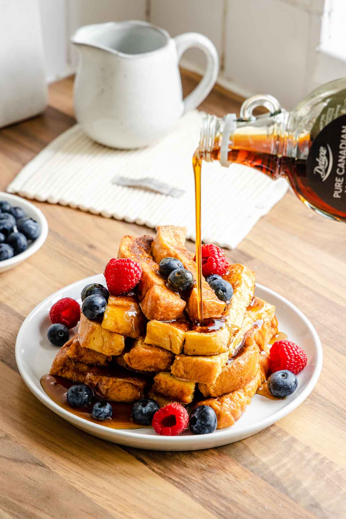 French Toast Sticks finished, stacked on a plate with berries, pouring syrup over it