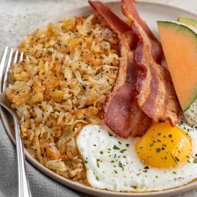 plate of crispy hash browns next to two pieces of bacon, a fried egg, and slices of cantaloupe.