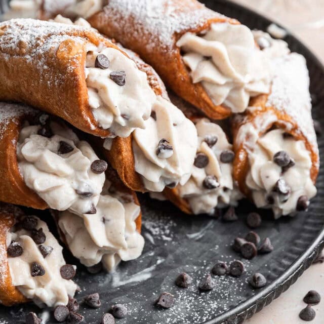 Cannoli Cream Filling prepared Cannolis stacked on black plate with chocolate chips on and around Cannolis. 4x3